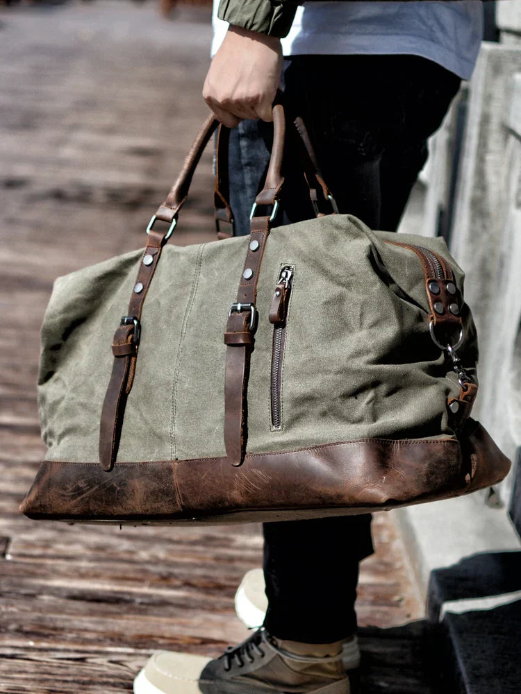 Person holding a large green duffel bag with brown leather straps on a wooden surface.